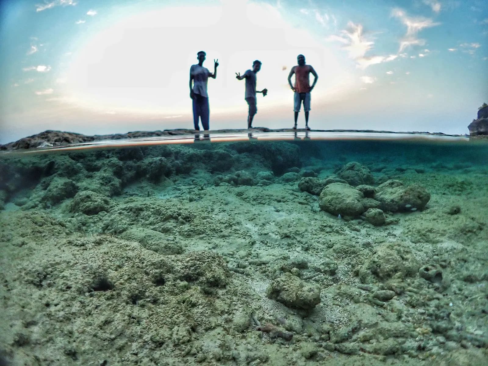 Wide over-under split shot of friends on rocky shore above a clear shallow reef