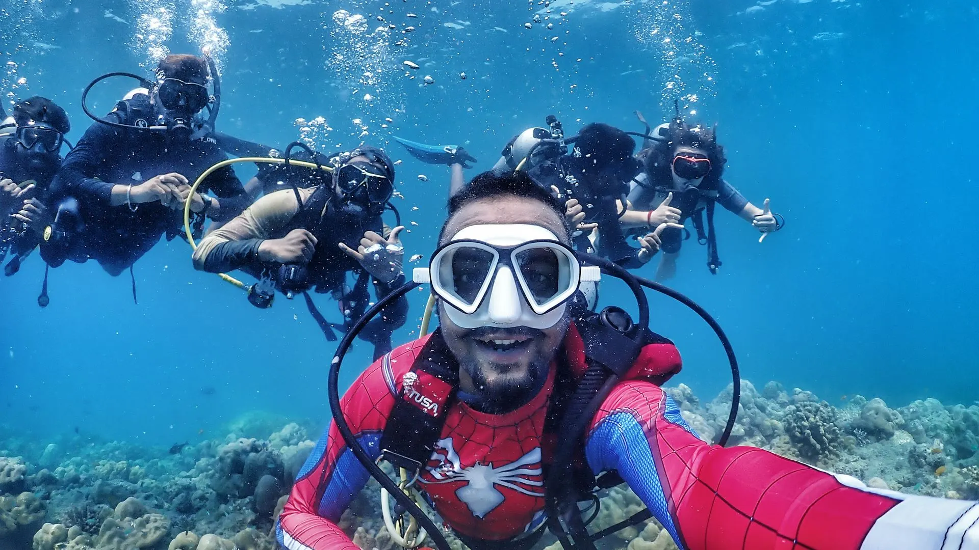 Oceanspidey in a Spider-Man dive suit taking an underwater group selfie with four divers over a Havelock reef
